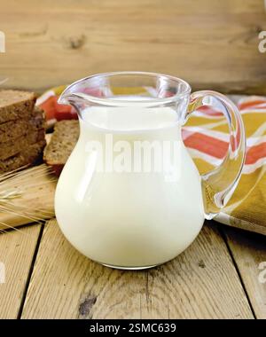 Milch in ein Glas Glas mit Roggenbrot, braun-rot karierte Serviette, Roggen Spike auf dem Hintergrund der Holzbretter Stockfoto