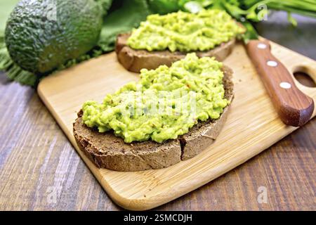 Zwei Scheiben Roggenbrot mit Guakomole-Avocado auf einem Teller, Serviette, Petersilie auf einem Holzbrett Hintergrund Stockfoto
