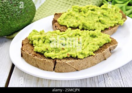 Zwei Scheiben Roggenbrot mit Guakomole-Avocado auf einem Teller, Serviette, Petersilie auf dem Hintergrund helle Holzbretter Stockfoto
