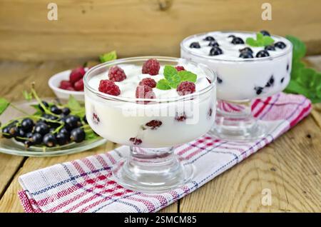 Dicken Joghurt in zwei Gläser mit Himbeeren und schwarzen Johannisbeeren auf eine Serviette, Beeren und Minze auf Holzbrettern Hintergrund Stockfoto