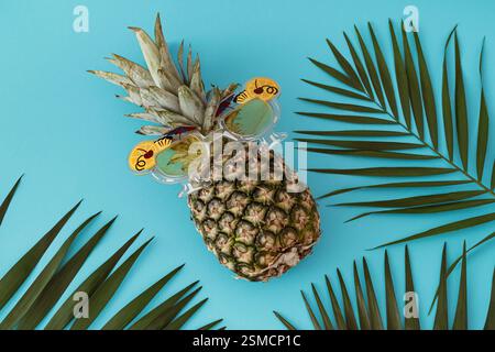 Sommerszene mit Ananas mit Party-Sonnenbrille und grünen Palmenblättern auf hellblauem Hintergrund. Minimales tropisches Konzept. Stockfoto