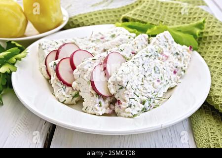 In Scheiben geschnittene Quark-Terrine mit Dill und Radieschen, grüne Zwiebeln, Salat auf einem Teller, gekochte Kartoffeln, Serviette, Petersilie auf hellen Holzdielen Stockfoto