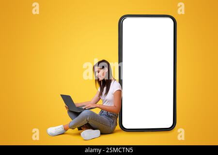 Lächelnde Frau, die einen Laptop benutzt und sich auf einen weißen, leeren Telefonbildschirm stützt Stockfoto