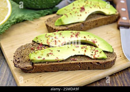 Zwei Scheiben Graubrot mit Scheiben von Avocado und Pfeffer, Zitrone, Serviette, Messer gegen die dunkle Holzplatte Stockfoto
