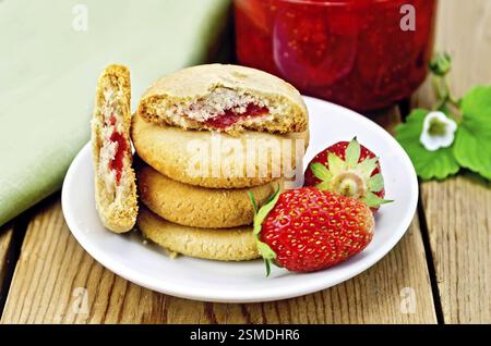 Einen Stapel von Cookies gefüllt mit Marmelade und Erdbeeren auf einem Teller, ein Glas Strawberry jam auf dem Hintergrund der Holzbretter Stockfoto