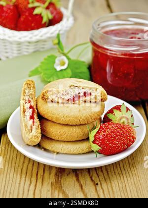Stapel von Cookies gefüllt mit Marmelade und Erdbeeren auf einem Teller, ein Glas Erdbeermarmelade, ein Weidenkorb mit einer Erdbeere auf Holzbrett Stockfoto
