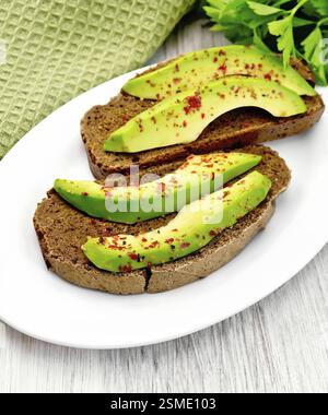 Zwei Scheiben Roggenbrot mit Scheiben Avocado und Pfeffer auf einem Teller, Serviette, Petersilie auf dem Hintergrund helle Holzbretter Stockfoto