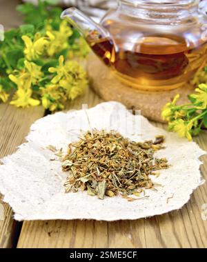 Getrocknete Blüten von Hypericum auf einem Stück Papier, Tee in der Glas-Teekanne und frische Blumen Tutsan auf dem Hintergrund von Holzbrettern Stockfoto