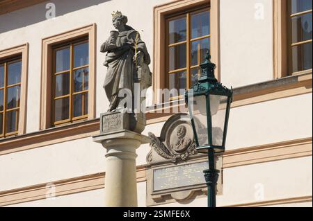 Steinstatue eines heiligen mit Kelch und Kreuz, stehend auf einer Säule. Hintergrund mit Fenstern und grünem Laternenpfahl. Tschechische Republik. Stockfoto