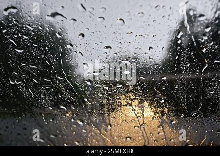 Regentropfen auf Glasfenster mit verschwommenen Bäumen, der Straße und den gelben Lichtern des Autos Stockfoto