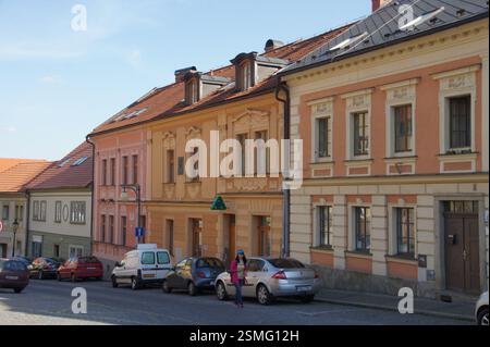 Straßenszene in Tschechien. Farbenfrohe Gebäude mit kunstvollen Fassaden säumen eine kopfsteingepflasterte Straße. Autos parkten am Bordstein. Eine Frau in einer rosa Jacke Stockfoto