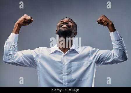 Studio-Porträt eines jungen afrikanischen Geschäftsmannes, der seinen Bizeps beugt und mit einem großen Lächeln aufblickt und in seinem weißen Hemd Selbstvertrauen und Kraft ausdrückt Stockfoto