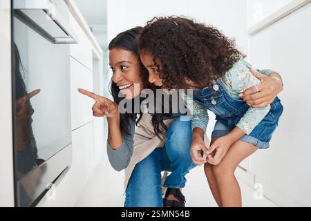 Mutter, Tochter und Backen im Ofen in der Küche zum Warten, zeigen auf Kuchen und Timing Dessert. Lernen, Mädchen und neugierig am Herd Stockfoto