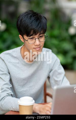 Ein junger asiatischer Mann arbeitet von einem Café aus ferngesteuert an seinem Laptop und sitzt an einem Tisch im Freien. Menschen, drahtlose Technologie Stockfoto