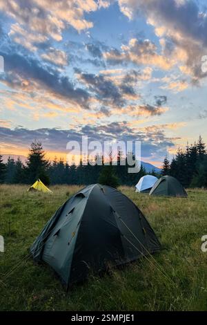 Wenige Touristenzelte in einem grasbewachsenen Feld am Abend, umgeben von Bäumen und Hügeln unter atemberaubendem Sonnenuntergangshimmel. Farbenfrohe Wolken verleihen der Szene eine schöne Note mit leuchtenden Orange- und Violetttönen am Horizont. Stockfoto