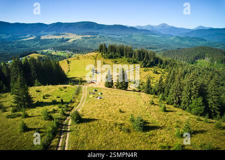 Campingplatz aus der Vogelperspektive in den Bergen, mit mehreren bunten Zelten auf einer grasbewachsenen Rodung. Camper entspannen unter klarem blauen Himmel, umgeben von hohen immergrünen und Panoramablick auf entfernte Gipfel. Stockfoto