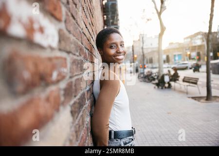 Stilvolle junge afroamerikanische Frau mit kurzen Haaren, die lächelnd an eine Ziegelmauer in einer Stadtstraße lehnt Stockfoto