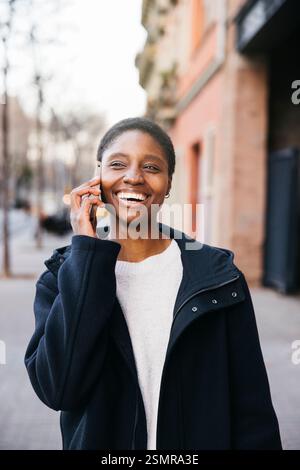 Fröhliche junge Frau, die ein angenehmes Telefongespräch führt, während sie die Straße hinunter läuft Stockfoto