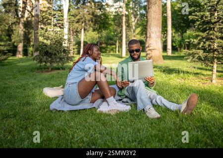 Ein ruhiges afroamerikanisches Paar sitzt unter Bäumen und lernt online. Man durchsucht die Prüfungsressourcen auf Gadget Stockfoto