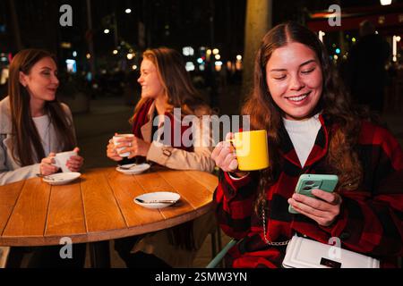Drei Junge Frauen Sitzen Zusammen Und Schlürfen Ihren Kaffee, Während Sie Im Gespräch Sind. Eine Frau lächelt, während sie ein Smartphone überprüft. Ihre Freudige Präsenz Unterstreicht Freundschaft Und Verbundenheit. Hochwertige Fotos Stockfoto