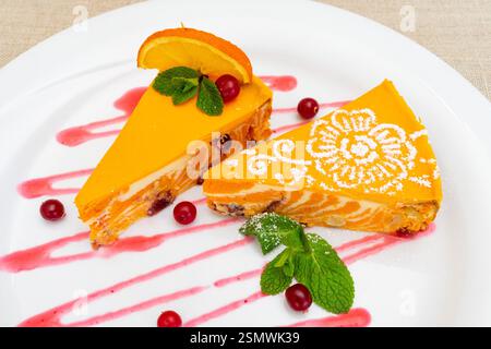 Zwei Scheiben Mango Mousse Dessert garniert mit Minzblättern, Preiselbeeren und Orangenscheiben werden künstlerisch auf einem weißen Teller präsentiert Stockfoto