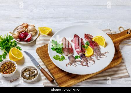 Frischer roher Tintenfisch mit Zitrone und Petersilie auf weißem Teller auf Schneidebrett mit Gewürzen, Serviette und Messer auf weißem Holztisch, horizontale Ansicht Stockfoto