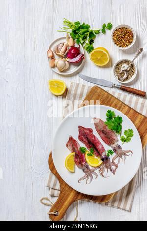 Frischer roher Tintenfisch mit Zitrone und Petersilie auf weißem Teller auf Schneidebrett mit Gewürzen, Serviette und Messer auf weißem Holztisch, horizontale Ansicht Stockfoto