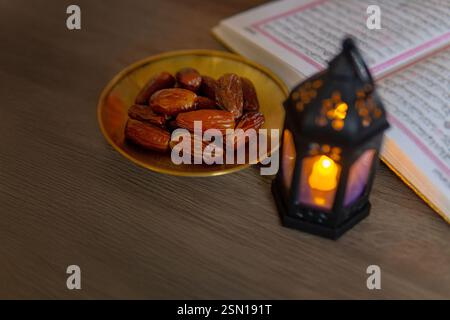 Eine Schüssel Datteln, eine beleuchtete Laterne und ein offener Al-Quran auf einem Holztisch, symbolisiert den islamischen heiligen Monat Ramadan. Kopierbereich für Text. Stockfoto