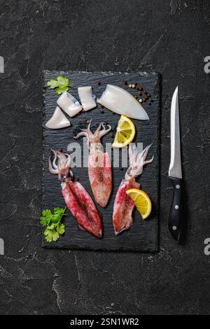 Frischer roher Babykalmar mit Zitrone und Petersilie auf schwarzem Schieferteller mit Messer auf Betontisch, horizontaler Blick von oben, flache Lage Stockfoto