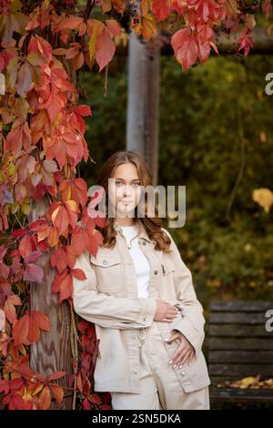 Herbstporträt: Stilvolle junge Frau in Beige Outfit posiert von Holzpergola mit roten Herbstblättern. Gemütliches Herbstporträt in landschaftlich reizvollem Garten Stockfoto