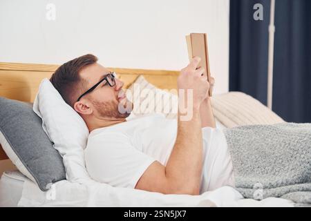 Mann in weißem Hemd und Brille, der auf dem Bett liegt und Buch liest. Stockfoto