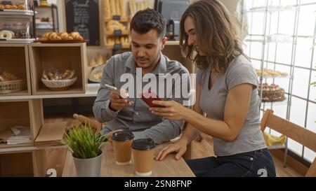 Ein Paar sitzt zusammen in einer gemütlichen Bäckerei, genießt Kaffee und nutzt sein Smartphone, während es von frischem Gebäck umgeben ist Stockfoto