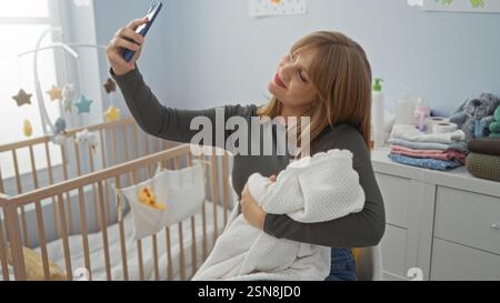 Eine Frau, die im Kinderzimmer Selfie macht, das Baby in eine Decke gewickelt hält, umgeben von Kinderbett, Plüschspielzeug und Babyzubehör, hält einen fröhlichen Moment in der Wohnung fest. Stockfoto