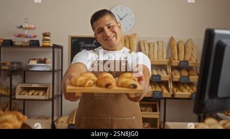 Junger hispanischer Mann, der in einer Bäckerei arbeitet, lächelt und ein Tablett mit Croissants hält, während er in einem ordentlich organisierten Brotladen mit Regalen steht Stockfoto