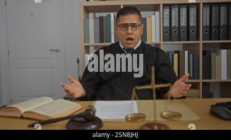 Der junge hispanische Mann in einem Richtergewand sieht überrascht aus in einem Gerichtssaal mit Rechtsbüchern, Justizmaßstäben und einem Hammer auf dem Schreibtisch. Stockfoto