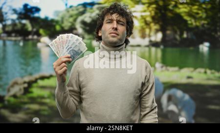 Ein spanischer Mann, der polnische Zloty-Banknoten hält, steht selbstbewusst in einem üppigen Park, umgeben von lebhaften grünen Bäumen mit einem ruhigen Teich im Hintergrund Stockfoto