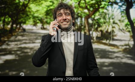 Junger Mann in einem Park, der am Telefon telefoniert, elegant gekleidet von Bäumen an einem sonnigen Tag, schafft eine fesselnde und professionelle Atmosphäre mit Natur Stockfoto
