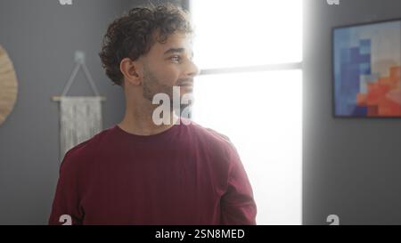 Ein junger Mann mit lockigen Haaren steht drinnen in einem gut beleuchteten Wohnzimmer und bietet eine entspannte, aber moderne Atmosphäre in seinem stilvollen Apartment. Stockfoto
