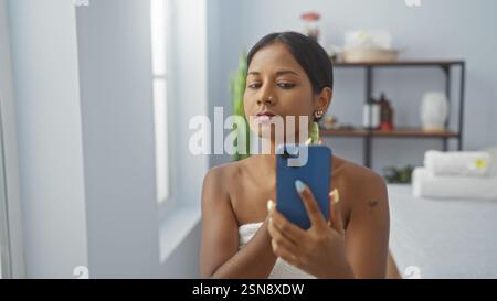 Eine junge Frau in einem Spa-Raum, die mit ihrem Smartphone ein Selfie macht, während sie in ein Handtuch gewickelt ist und einen Moment der Entspannung und Selbstpflege in einer ruhigen Atmosphäre festhält Stockfoto
