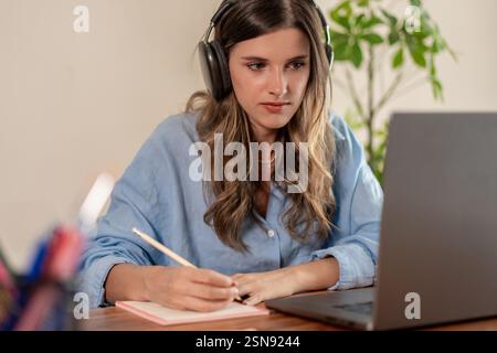 Junge Frau in blauem Hemd, die Kopfhörer trägt und Notizen schreibt, während sie zu Hause an ihrem Laptop arbeitet. Stockfoto