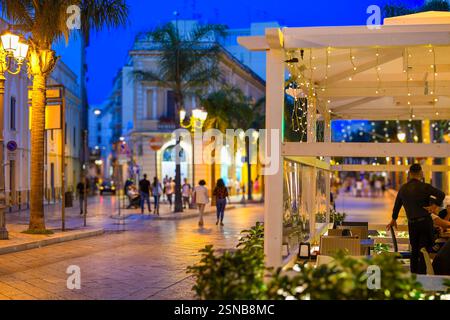 Ein männlicher Kellner wartet unter einem Portikus in einem Straßencafé an der Hauptstraße Corso Umberto zur dämmerblauen Stunde in Brindisi Italien, Apulien auf Kunden Stockfoto