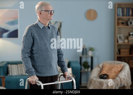 Mittlere vollständige Aufnahme eines ruhigen älteren Mannes, der langsam im Wohnzimmer läuft und Rahmen hält, während er sich im Haus, im Kopierraum bewegt Stockfoto
