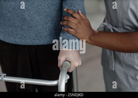 Zugeschnittene Aufnahme einer nicht erkennbaren schwarzen Sozialarbeiterin, die den Arm des Mannes berührt und ältere Patienten beim langsamen Gehen mit dem Rahmen unterstützt, während sie sich im Haus bewegen, Konzept für betreutes Wohnen Stockfoto