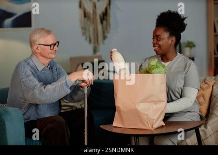 Mittelgroße Aufnahme einer schwarzen Pflegerin, die Milchprodukte und frisches Gemüse an einen einsamen älteren Mann liefert, um älteren Menschen beim Einkaufen von Lebensmitteln im Wohnzimmer zu helfen Stockfoto