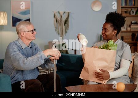 Lächelnde schwarze Sozialarbeiterin liefert Milchprodukte und frisches Gemüse an ältere Menschen mit Behinderung und hilft älteren Menschen beim Einkaufen von Lebensmitteln, während sie auf der Couch im Wohnzimmer sitzt Stockfoto