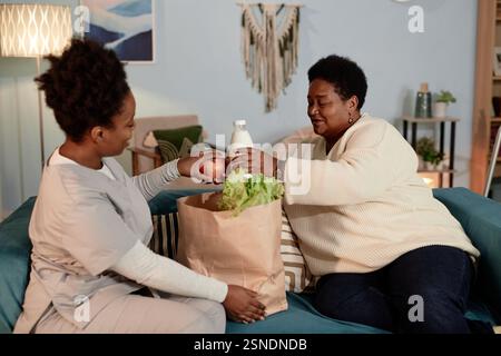 Mittelgroße Aufnahme in warmen Tönen einer zufriedenen schwarzen Seniorin, die Papiertüte mit Lebensmitteln von Sozialarbeiterinnen erhält, um älteren Menschen bei der Lieferung von Lebensmitteln zu Hause zu helfen Stockfoto