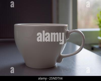 Porzellan-Kaffeetasse in weiß, dampfend in der Nähe von sonnendurchfluteten Fenstern, warmes Licht über die Holzoberfläche mit sanfter Morgenbeleuchtung Stockfoto