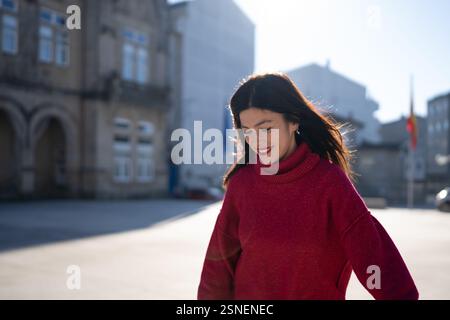 Glückliche Frau in einem leuchtend roten Pullover und dazu passendem Lippenstift, der durch einen belebten Marktplatz in der Innenstadt spaziert und die Wärme eines sonnigen Wintertages aufnimmt Stockfoto