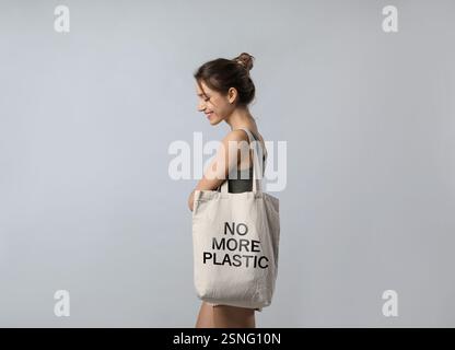 Glückliche junge Frau, die eine Öko-Tasche mit No More Plastik-Text auf hellgrauem Hintergrund hält Stockfoto