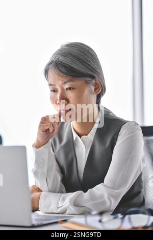 Geschäftige, ernsthafte asiatische Geschäftsfrau mittleren Alters, die im Büro mit einem Laptop arbeitet. Stockfoto